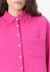 Camicia rosa, con bottoni, realizzata in tessuto testurizzato, dotata di colletto a punta e una tasca sul petto. Bottoni bianchi adornano la parte anteriore.