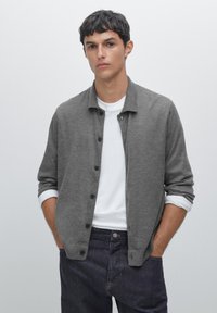 Grå stickad cardigan med knappfront, mjuk textur och krage, lagd över en vit t-shirt. Mörka jeans fullbordar outfiten.