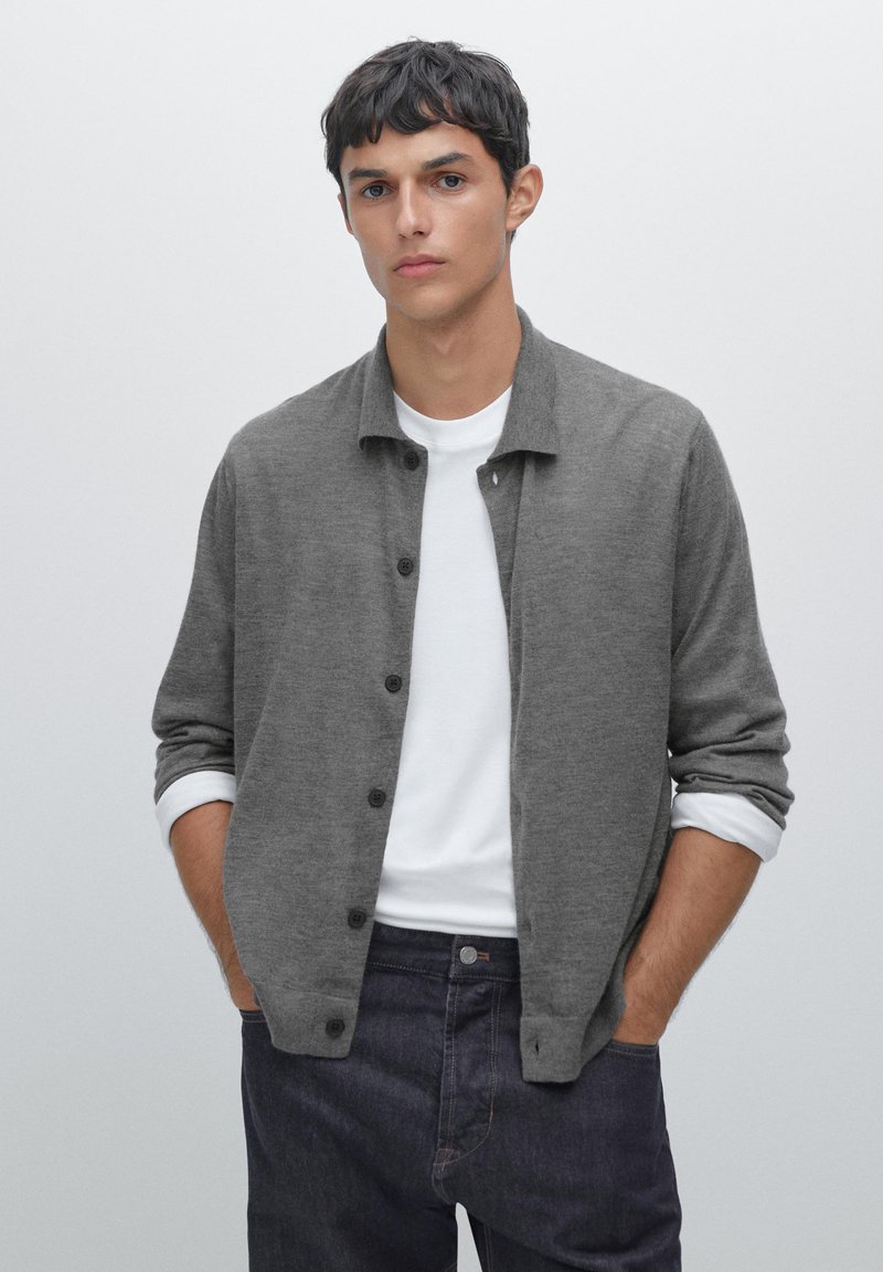 Grå stickad cardigan med knappfront, mjuk textur och krage, lagd över en vit t-shirt. Mörka jeans fullbordar outfiten.