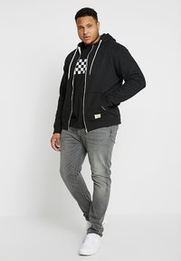 Čierna mikina na zips s bielymi šnúrkami, skombinovaná s čiernym tričkom s károvaným vzorom a šedými džínsami. Biely tenisky dopĺňajú outfit.
