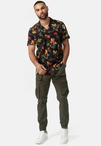 Chemise noire à fleurs à manches courtes avec des motifs vibrants orange et vert, associée à un pantalon cargo vert et des baskets blanches.