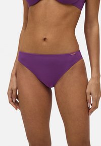 Parte inferior de bikini morada con una textura suave y un diseño de corte bajo, que presenta costuras mínimas y un pequeño logotipo de metal como acento en el lado.