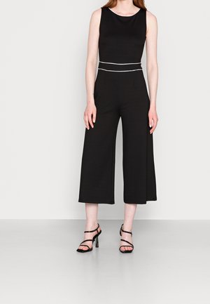 Kvinde iført ærmeløs sort jumpsuit med hvidt bælte og brede udsvingede bukser, kombineret med sorte remstiletter med høj hæl.