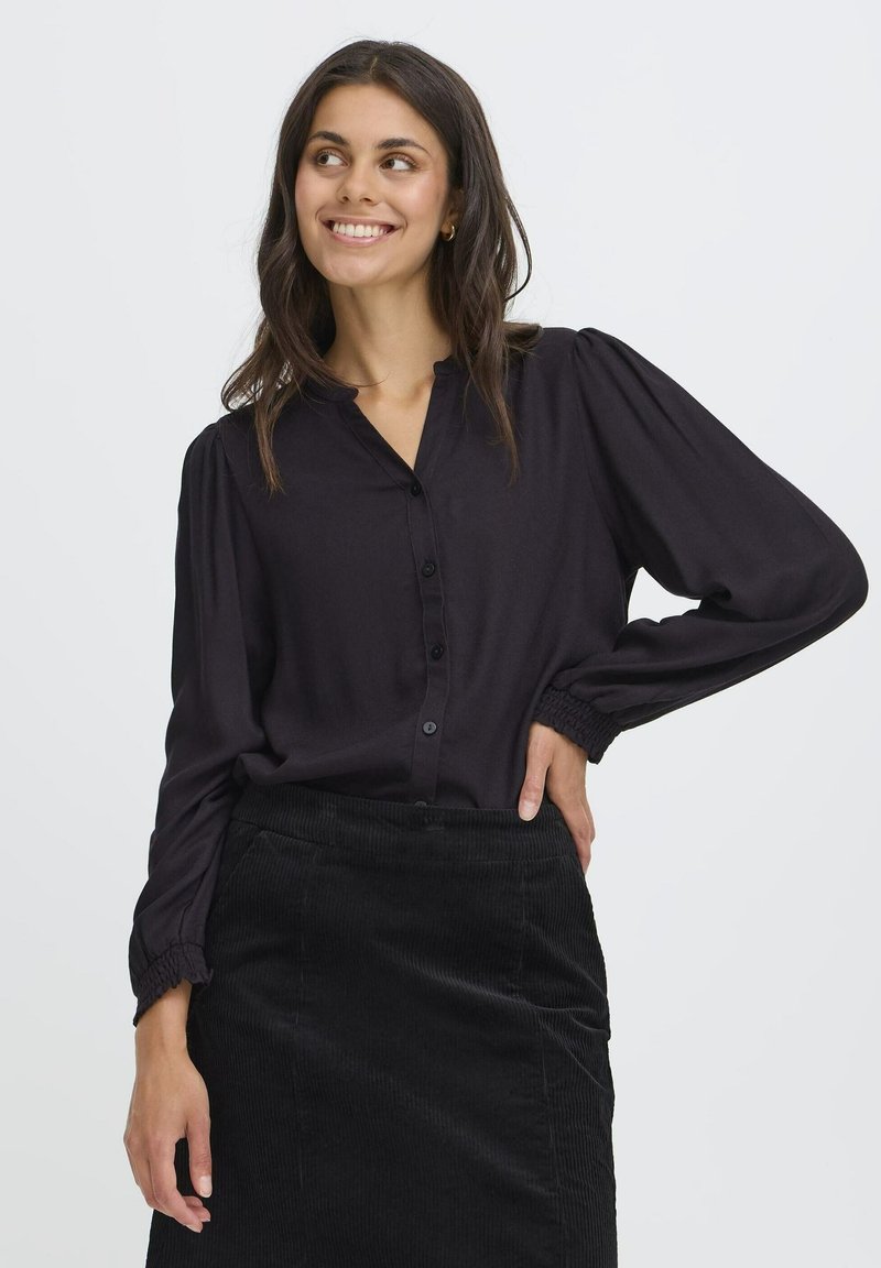 Blusa nera a maniche lunghe con bottoni frontali e polsini arricciati, abbinata a una gonna in corduroy nera. Tessuto liscio con leggero lucido.