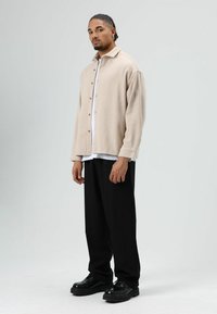 Beige overhemd met knopen, losse pasvorm, gedragen over een wit T-shirt. Zwarte broek met wijde pijpen en glanzende zwarte schoenen maken de look compleet. Glad textuur van de stof.