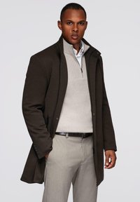 Abrigo marrón con un cuello alto, cierre de cremallera y bolsillos laterales, llevado sobre un suéter beige claro y pantalones grises. Tejido suave y estructurado.