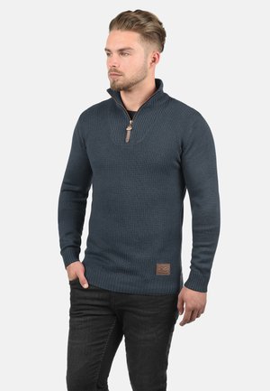 Homme portant un pull texturé bleu marine à demi-fermeture éclair avec poignets et ourlet côtelés, associé à un jean noir, debout avec une main dans la poche.
