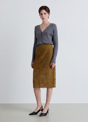 Donna in piedi che indossa un cardigan grigio a trecce con scollo a V, gonna di pizzo color senape alla lunghezza del ginocchio e décolleté nere con cinturino posteriore su sfondo semplice.