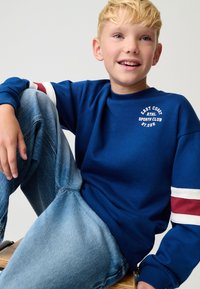 Felpa blu con strisce bianche e marroni sulle maniche, con stampa del logo. Indossata con jeans di denim chiaro, seduto in modo casual.