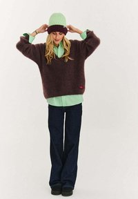 Přerostlý, chlupatý svetr švestkové barvy s výstřihem do V, vrstvený přes světle zelenou blůzu. Osoba má na sobě mátově zelenou čepici s tmavým lemem. Rozšířené tmavé džíny.