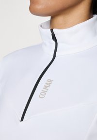 Bílý top s vysokým límcem na zip s černým zipem a logem "COLMAR" v béžové barvě na hrudi. Hladká textilie a obepínající design.