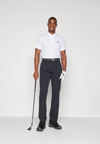 Hvit polo skjorte med logo, mørke, tettsittende bukser, svart belte, hvit golfhanske, holder en golfkølle, og svarte golfsko.