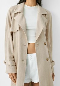 Beżowy trench z lekko fakturowanej tkaniny, z dużymi klapami i dwoma guzikami. Noszony na białej koronkowej bluzce i białych szortach.