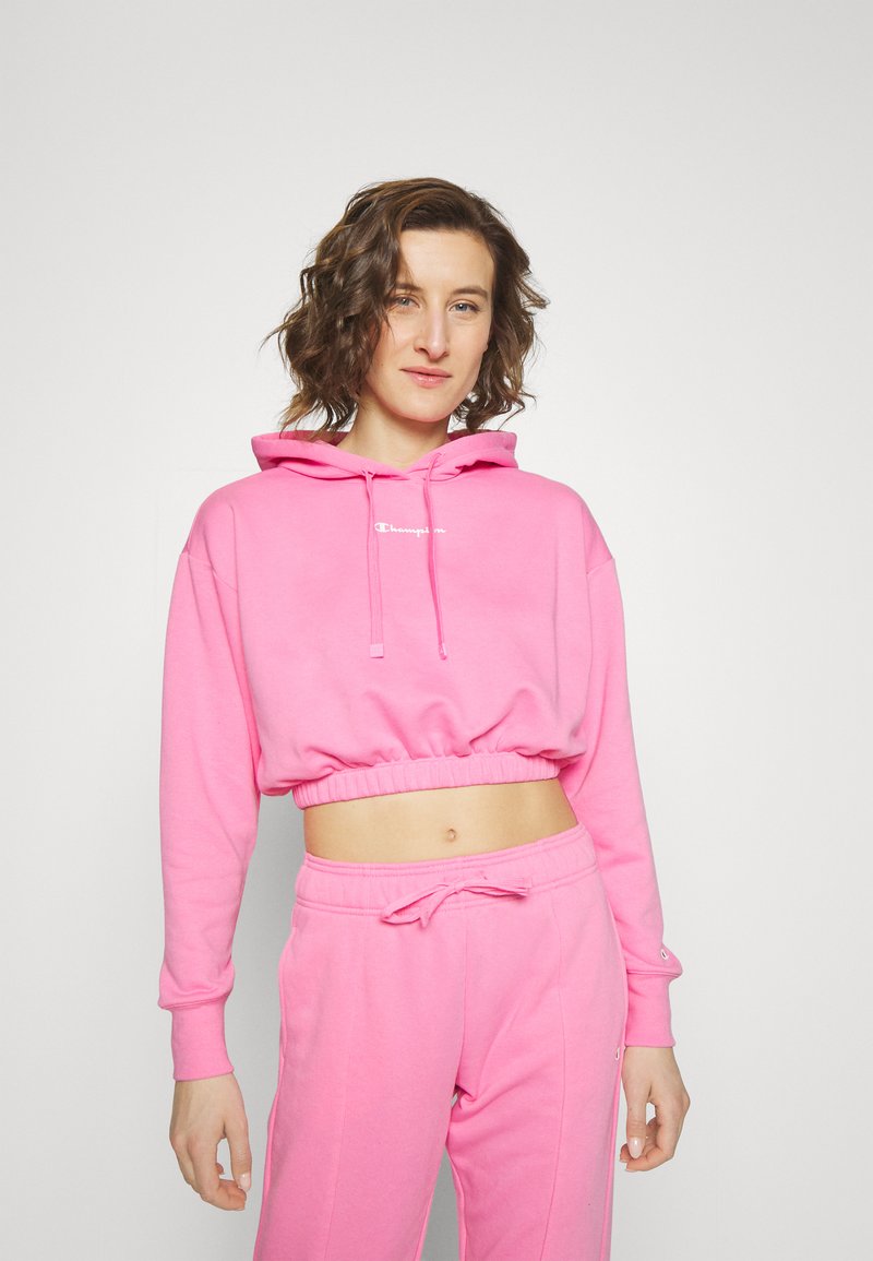 Felpa corta rosa con cappuccio, caratterizzata da un logo frontale e orlo a costine. Si abbina a pantaloni della tuta rosa coordinati, realizzati in un tessuto morbido.