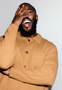 Beiger, gerippter Pullover mit Kragen, Knopfverschluss und zwei Fronttaschen. Das Model hat einen Bart und lacht, wobei es einen Teil seines Gesichts verdeckt.