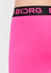 Lähedal olev pilt inimesest, kes kannab erksate roosa värvi leggings'eid, millel on must pael ja osaliselt nähtav roosa tekst.