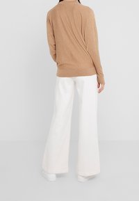Hellbrauner Strickpullover mit lockerer Passform und gerippten Bündchen, kombiniert mit weißen weiten Hosen und lässigen weißen Schuhen.