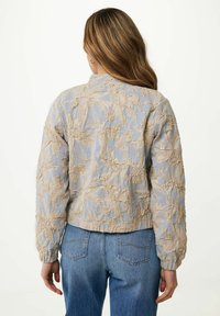 Veste bomber texturée en bleu clair avec broderie florale beige, manches et ourlet élastiques, portée avec un jean bleu taille haute.