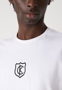 Biely bavlnený tričko s čiernym vyšitým logom v tvare štítu so znakmi "C" a "K" v strede.