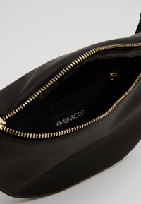 Sort faux læder taske med en glat tekstur, guld lynlåsdetaljer og et indvendigt mærke. Tasken har en rund form og en lynlåsåbning.