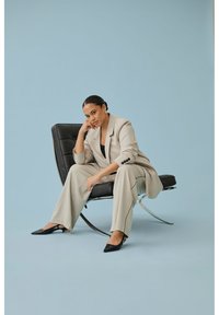 Frau im beigen Nadelstreifenanzug sitzt nachdenklich auf einem modernen schwarzen Ledersessel vor einem hellblauen Hintergrund.