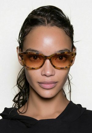 Femme aux cheveux mouillés portant des lunettes de soleil écaille avec des verres ambrés et un haut noir, faisant face à l'avant sur un fond uni.