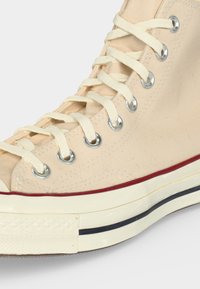 Κοντινό πλάνο ενός μπεζ καμβά ψηλού sneaker με λευκά κορδόνια, μεταλλικές οπές και κρεμ σόλα με κόκκινες και μαύρες ρίγες.