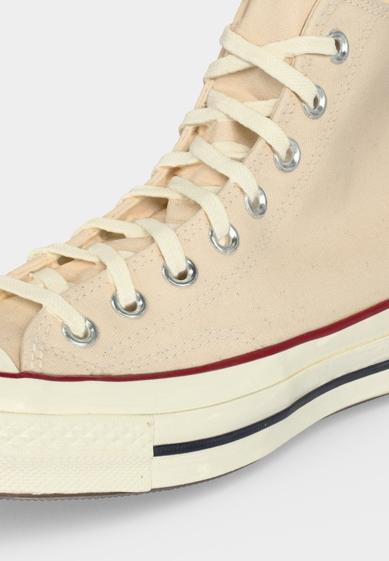 Nahaufnahme eines beigen High-Top-Sneakers aus Canvas mit weißen Schnürsenkeln, Metallösen und einer cremefarbenen Sohle mit roten und schwarzen Streifen.