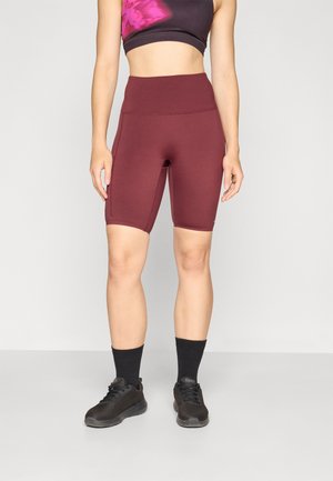 Shorts de vélo bourgogne à taille haute, fabriqués en tissu extensible. Associés à un crop top noir avec des accents tie-dye roses et des chaussures de sport noires.
