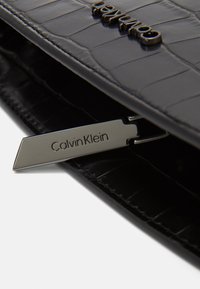 Czarna skórzana kopertówka o fakturze krokodyla, z metalową plakietką z wygrawerowanym napisem „Calvin Klein” oraz srebrnym detalem przypinanym.