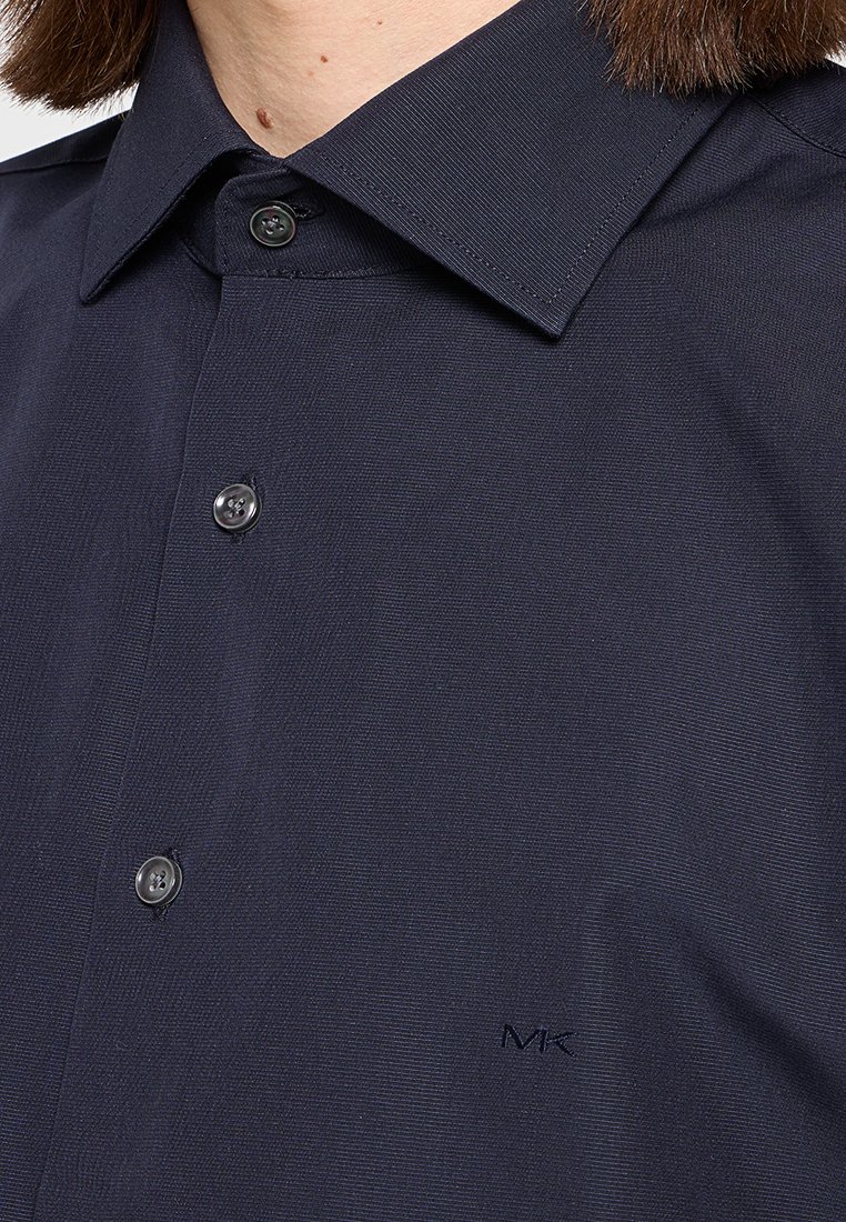 Camicia a maniche lunghe blu navy in tessuto testurizzato, con colletto a punta e piccolo logo sul petto. I bottoni neri sul fronte aggiungono dettagli.