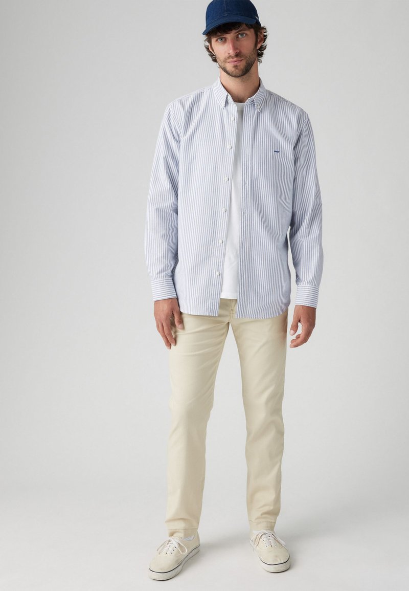 Camisa de botonadura azul claro y blanca a rayas, combinada con pantalones color crema y zapatillas blancas. Una gorra azul marino completa el conjunto.