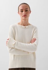 Cremefarvet strikket sweater med en struktureret ribkant ved halsen, der har dekorative knapper og en afslappet pasform. Lange ærmer med brede manchetter.