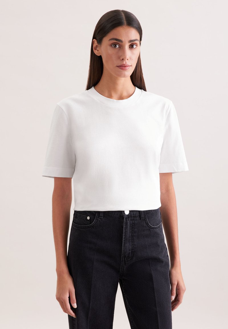 Femme aux cheveux longs et foncés portant un T-shirt blanc uni à manches courtes et un jean noir taille haute, sur un fond clair.
