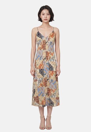 Donna con capelli corti e scuri che indossa un vestito midi patchwork con spalline sottili e tacchi aperti beige, in piedi su uno sfondo neutro.