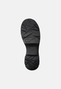 Suela de zapato de goma negra con un patrón de tracción en forma de cruz y un área central lisa con etiquetado en relieve. Diseño de punta redondeada.