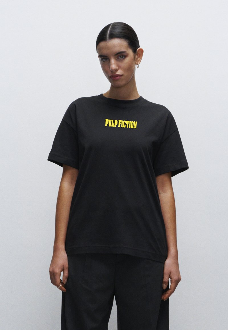 Svart t-shirt i bomull med korta ärmar, med gul text som står "PULP FICTION" över bröstet. En enkel design och passform.