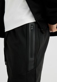 Pantalones deportivos negros con tejido texturizado, corte ajustado y bolsillos laterales con cremallera y detalles en negro. La cintura es elástica para mayor comodidad.