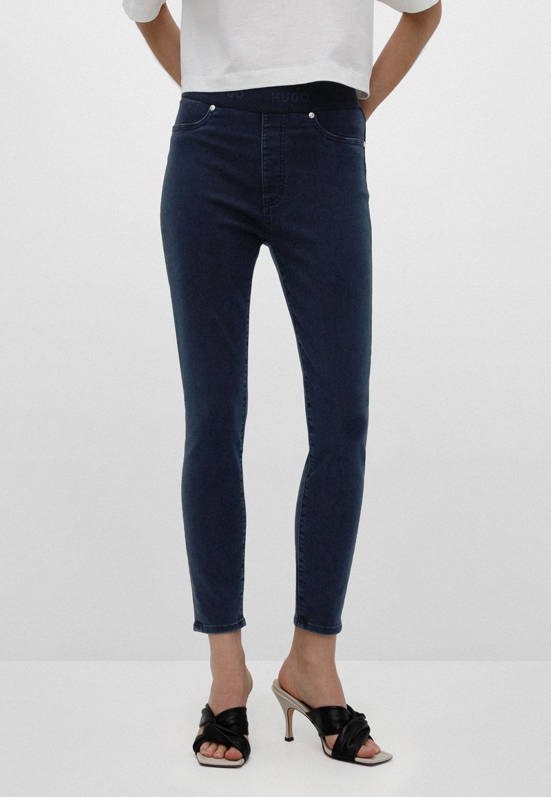 Personne portant un jean slim bleu foncé et des sandales à talons noirs avec des brides croisées, debout devant un fond uni.