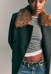 Giovane donna che indossa un cappotto scuro con colletto di pelliccia marrone, top grigio corto, jeans blu e una cintura nera con borchie, in piedi davanti a uno sfondo neutro.