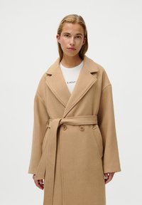 Cappotto avvolgente in misto lana beige con chiusura doppiopetto, ampi rever, grandi tasche e cintura da annodare. Texture morbida.