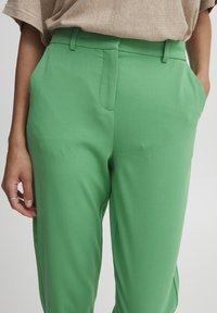 Pantaloni verdi su misura indossati da una persona con una camicia marrone chiaro, mani nelle tasche, concentrata sui dettagli dell'abbigliamento.