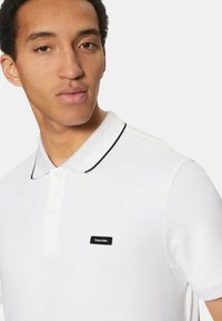 Polo bianca in cotone con finitura testurizzata, dotata di un colletto nero bordato e un piccolo logo Calvin Klein nero sul petto.