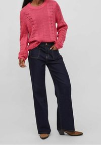 Maglione rosa lavorato a maglia con un motivo testurizzato e colletto rotondo abbinato a jeans a gamba larga blu scuro. I jeans presentano cuciture a contrasto.
