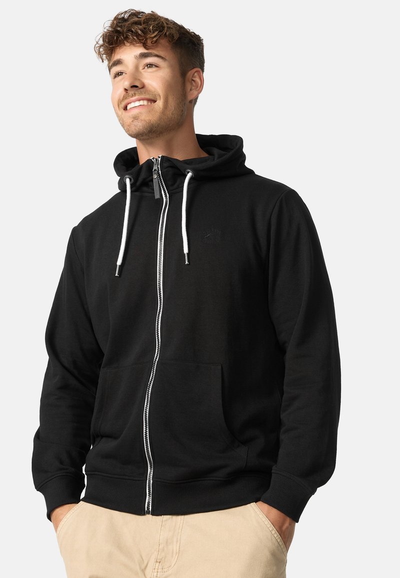 Zwarte zip-up hoodie van zacht materiaal. Heeft een capuchon, voorzakken, witte trekkoorden en een discreet logo op de borst.