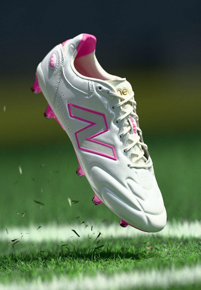 Chaussure de football New Balance blanc et rose en l'air au-dessus d'un terrain d'herbe avec des brins d'herbe éparpillés et des lignes blanches du terrain en dessous.