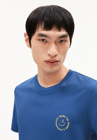 T-shirt in cotone blu con scollatura rotonda e maniche corte. Presenta un logo circolare dorato sul petto con testo e una faccina sorridente.