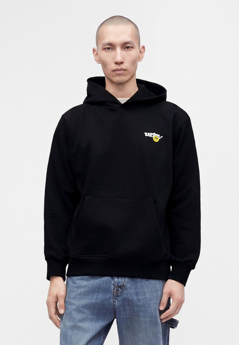 Homme portant un sweat à capuche noir avec un petit motif blanc et jaune sur la poitrine gauche et un jean bleu clair sur un fond uni.