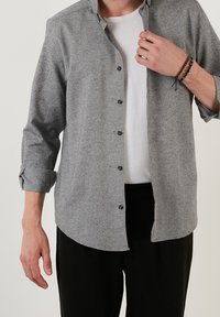 Homme portant une chemise grise à manches longues boutonnée sur un t-shirt blanc, pantalon noir, avec des bracelets au poignet droit, ajustant le col de la chemise.