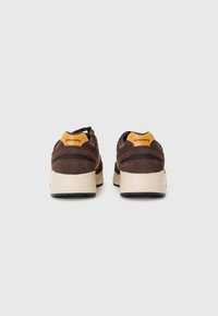 Baskets en daim marron avec un logo jaune contrastant au talon, comprenant des accents en mesh et une semelle en caoutchouc de couleur crème.